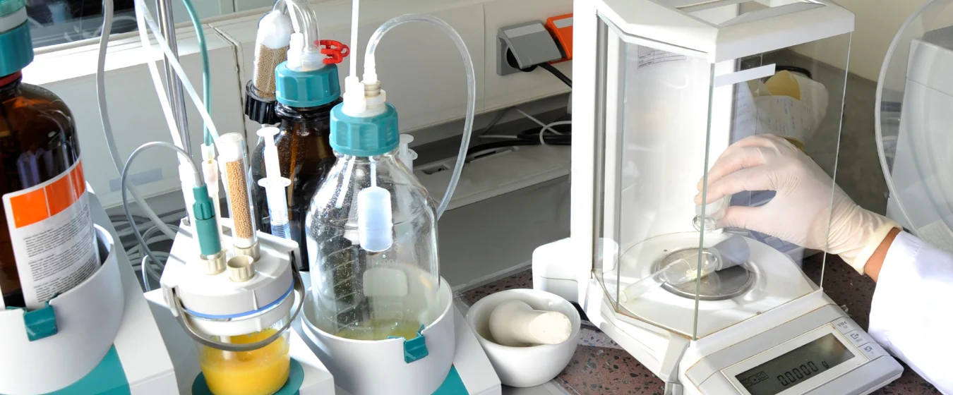 Titulador Karl Fischer com frascos de reagentes e eletrodo ao lado de balança analítica, onde técnico de luvas pesa amostra para medição de água em laboratório